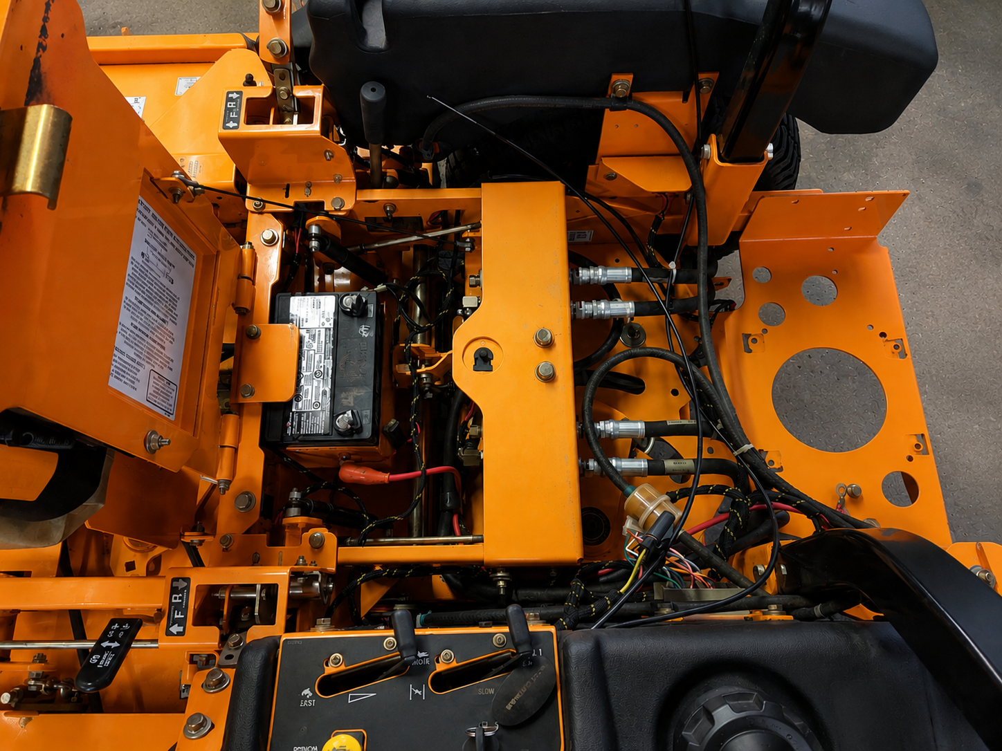Open equipment service deck showing battery connections, colored wiring harnesses, hydraulic hoses, and operator controls during diagnostics